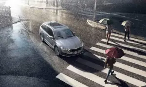 Srebrny kombi na mokrej ulicy podczas ulewy. Wycieraczki pracują, bo czujnik deszczu w samochodzie wykrył opady. Ludzie z parasolami przechodzą przez pasy.