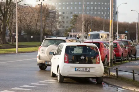 Biały Volkswagen up! i inne samochody stoją na pasach dla pieszych, blokując skrzyżowanie.