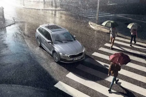 Srebrny kombi na mokrej ulicy podczas ulewy. Wycieraczki pracują, bo czujnik deszczu w samochodzie wykrył opady. Ludzie z parasolami przechodzą przez pasy.