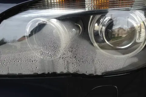 Parujące reflektory LED w nowym aucie, pokryte drobnymi kropelkami wody. Widać odbicie domu w soczewce.