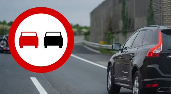 Znak zakazu wyprzedzania. Czerwony okrąg z dwoma samochodami, jeden czerwony, drugi czarny. Na drodze czarny SUV.