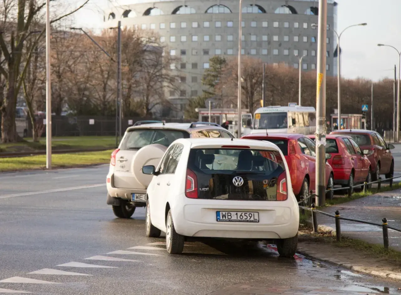 Biały Volkswagen up! i inne samochody stoją na pasach dla pieszych, blokując skrzyżowanie.