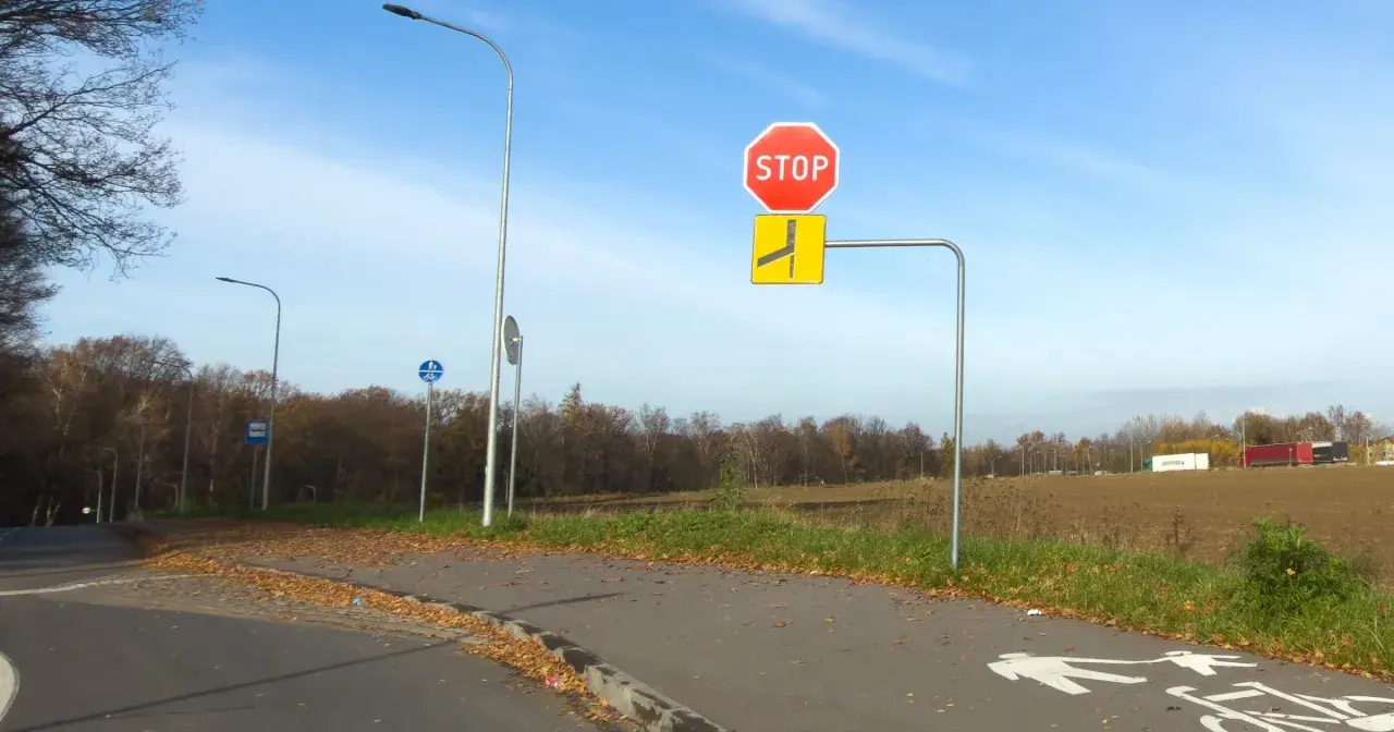 Znak STOP i znak informujący o łamanym pierwszeństwie po lewej stronie drogi. Jesień, liście na ścieżce rowerowej.