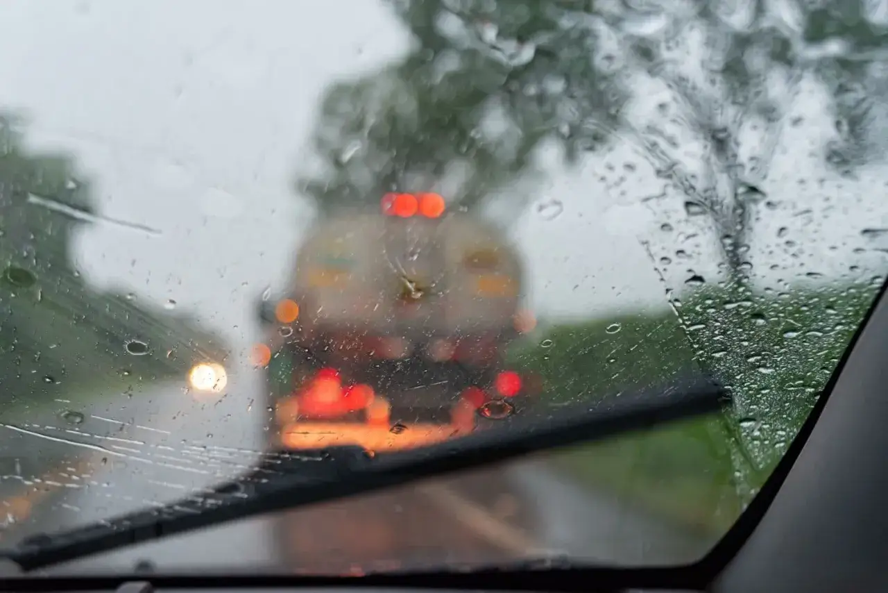 Widok przez zaparowaną szybę samochodu podczas deszczu. Wycieraczka pracuje, a przed nami widać światła innych pojazdów. Czujnik deszczu w samochodzie działa.