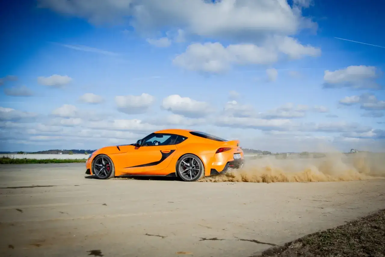 Pomarańczowe auto sportowe zrywa piasek podczas startu z launch control. Dynamiczna jazda pod błękitnym niebem.