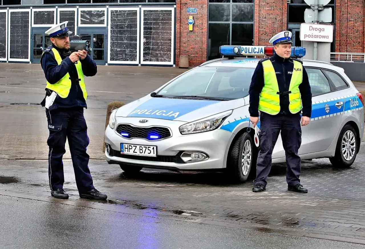 Policjanci z radarem mierzą prędkość. Jeden z nich celuje w kierunku drogi, drugi stoi obok radiowozu. Może to być kontrola przekroczenia prędkości o 15 km/h.
