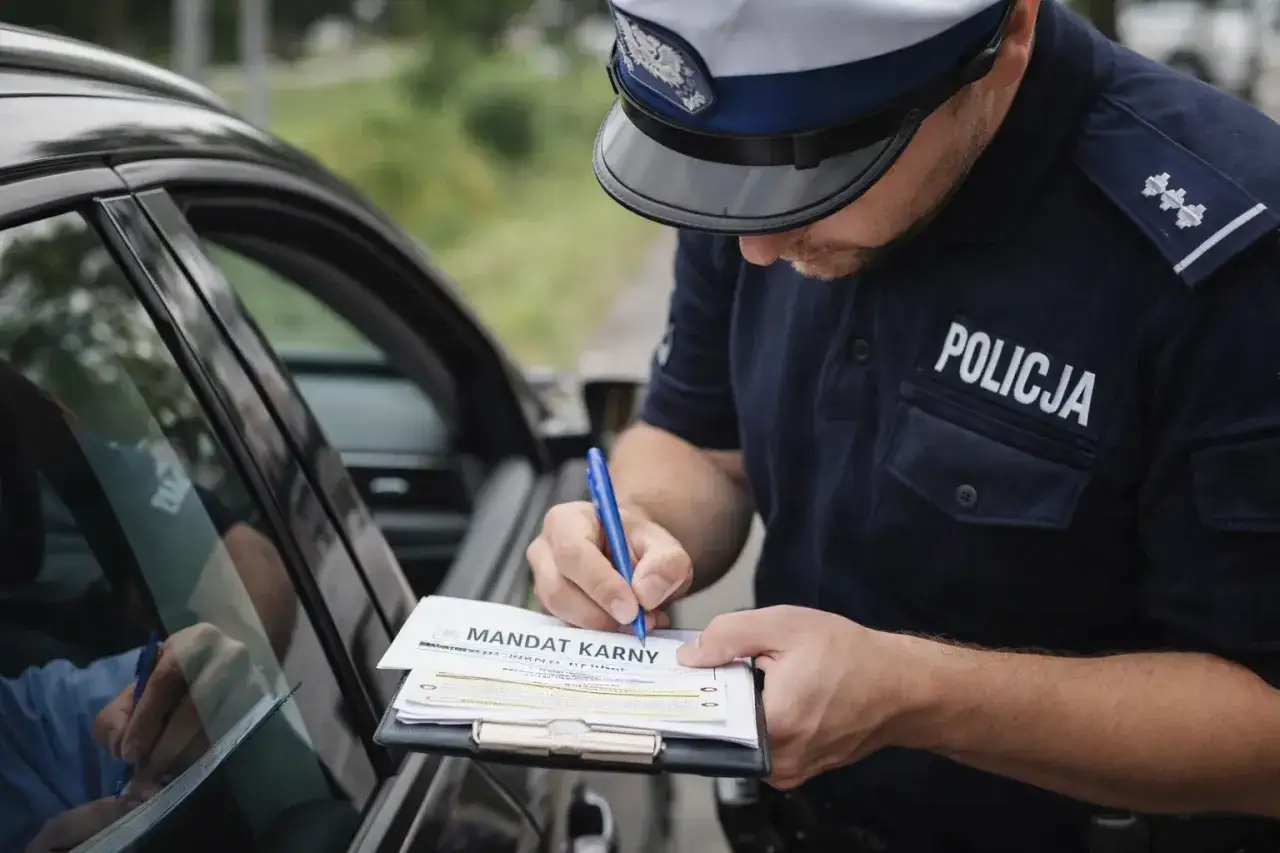 Policjant wypisuje mandat za przekroczenie prędkości o 20 km/h. Kierowca siedzi w samochodzie i podpisuje dokument.