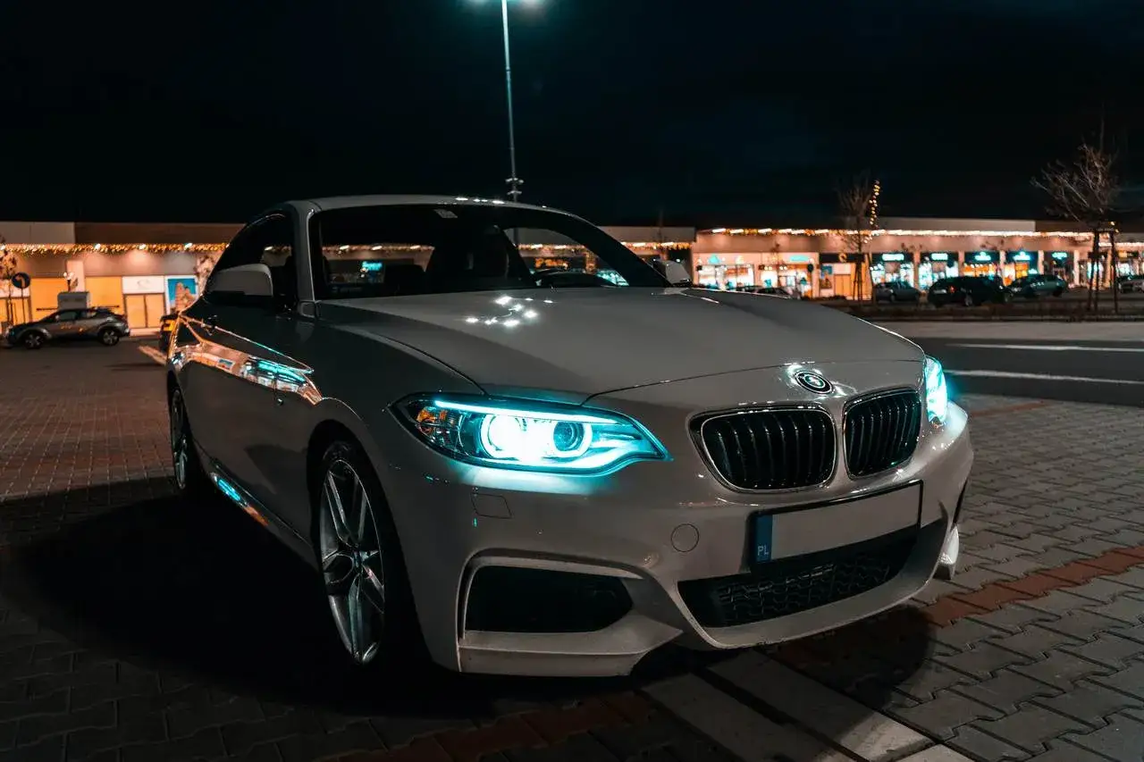 Białe BMW z włączonymi niebieskimi światłami AFS na nocnym parkingu. W tle widać oświetlone sklepy.