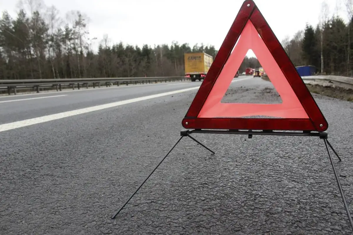 Czerwony tr&oacute;jkąt ostrzegawczy odległości stoi na poboczu drogi, sygnalizując potencjalne niebezpieczeństwo. W tle widać jadące samochody i drzewa.