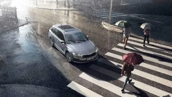 Srebrny kombi na mokrej ulicy podczas ulewy. Wycieraczki pracują, bo czujnik deszczu w samochodzie wykrył opady. Ludzie z parasolami przechodzą przez pasy.